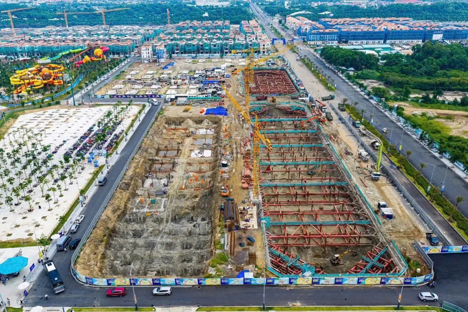 Ảnh chụp công trường một phân khu cao tầng Blanca City tháng 07/2026, hố móng lớn với hệ cốp pha đỏ và cần cẩu tháp, bên trái là công viên nước đã vận hành