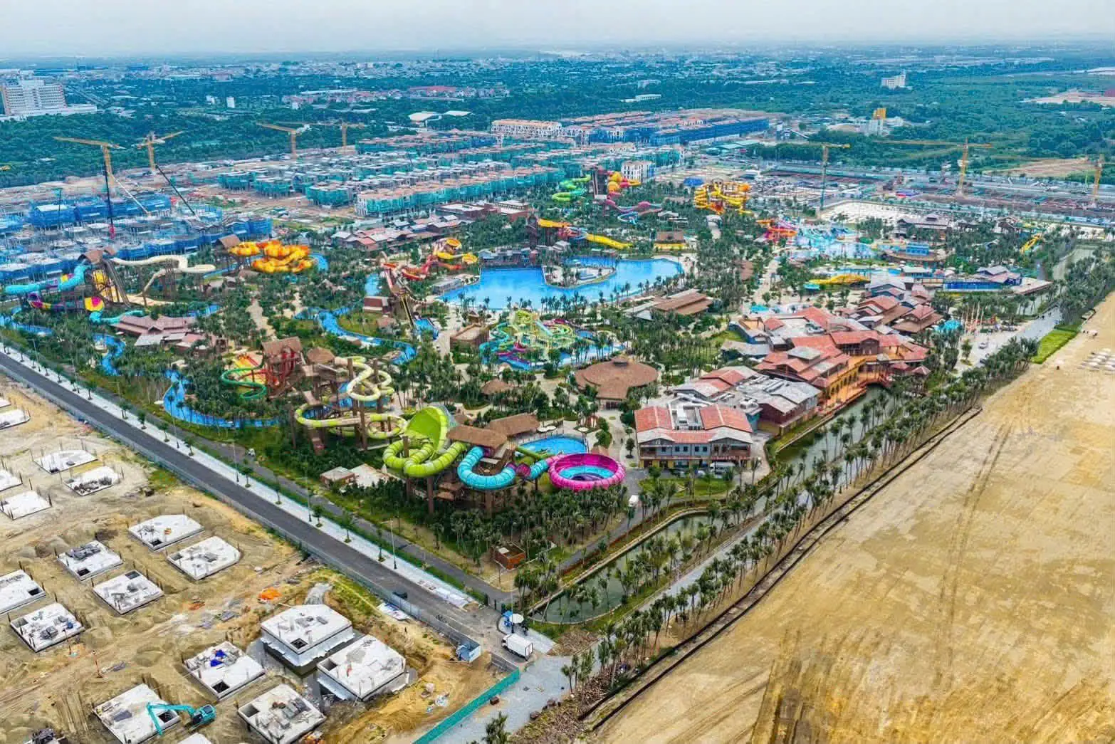Ảnh chụp công viên nước Sun World trong Blanca City tháng 07/2026 đang vận hành với các đường trượt và hồ tạo sóng, phía trước là các đài móng rời rạc của dải nhà phía biển